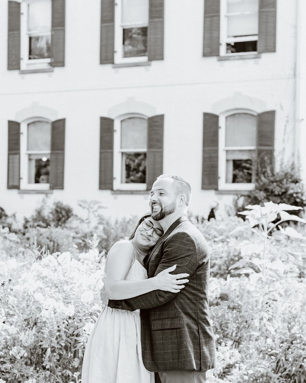 Black and white photo of a couple embracing in front of a white building with shuttered windows.