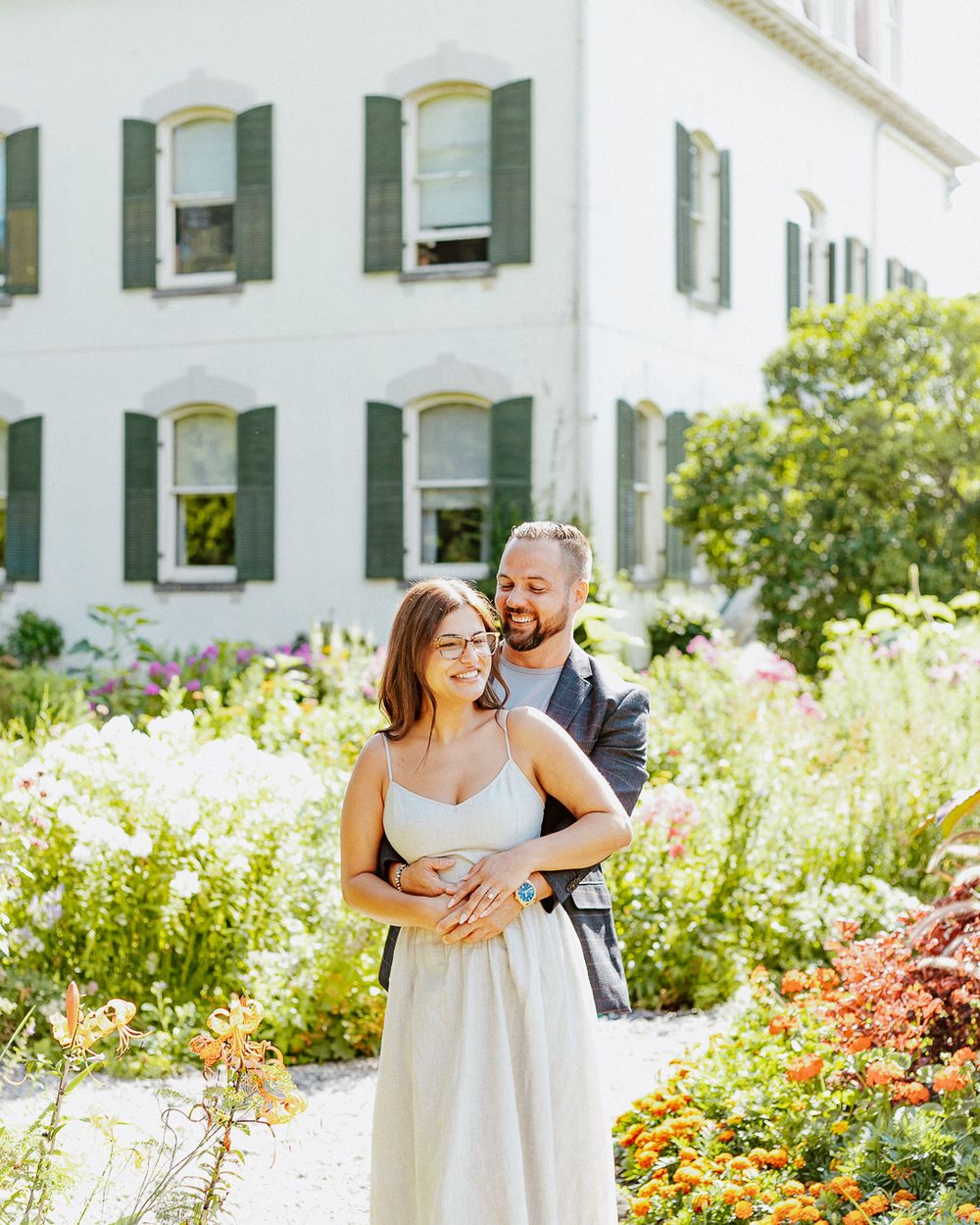 Couple embracing in a sunny garden with blooming flowers and a white house with green shutters.