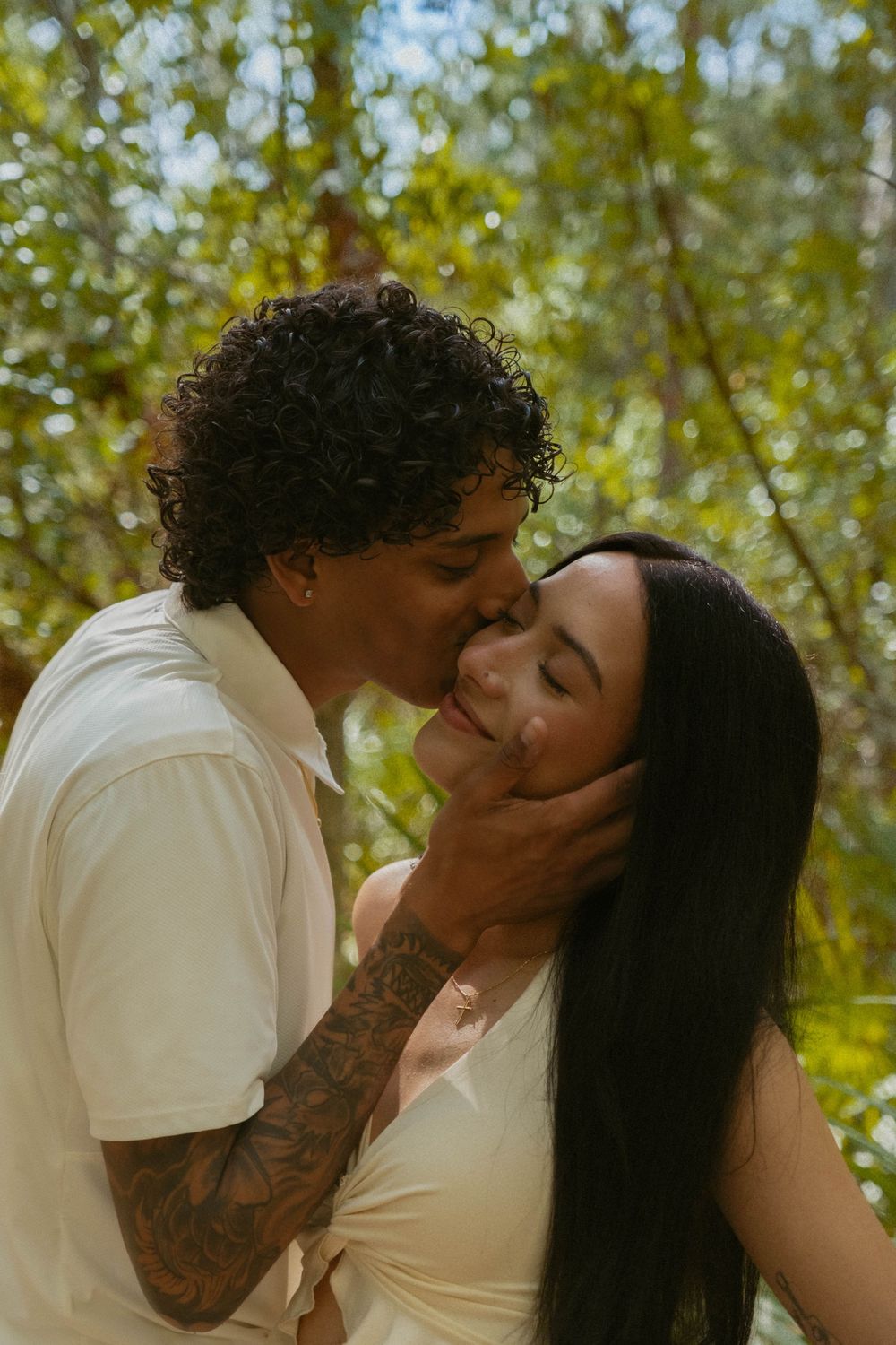 A tender moment shared in a sun-dappled outdoor setting with lush greenery.