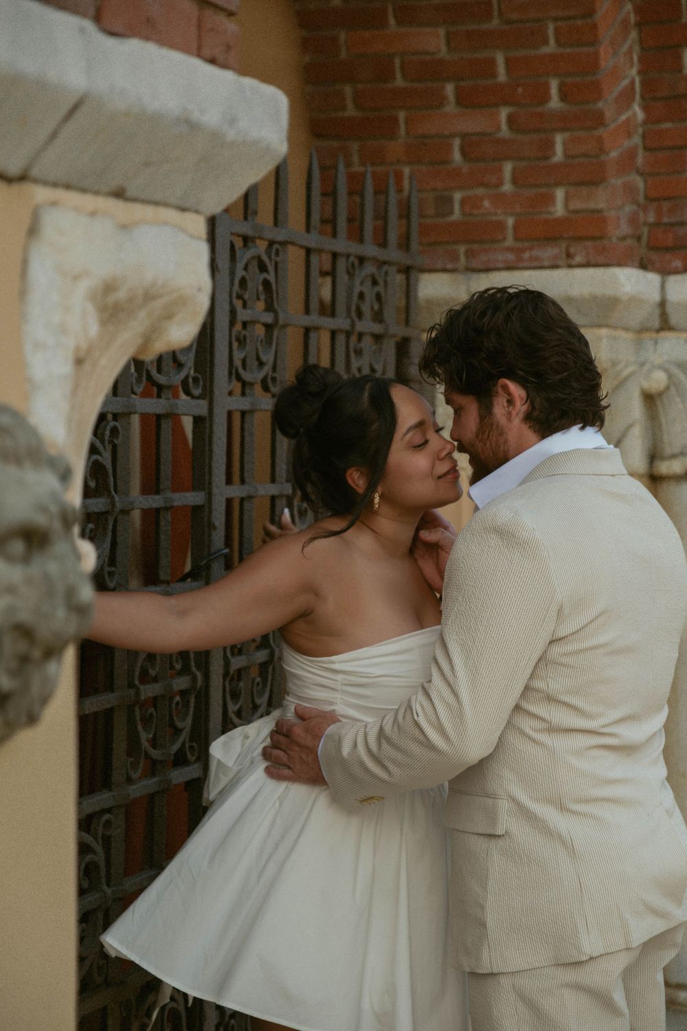 A couple in wedding attire shares a tender embrace against a brick wall backdrop.