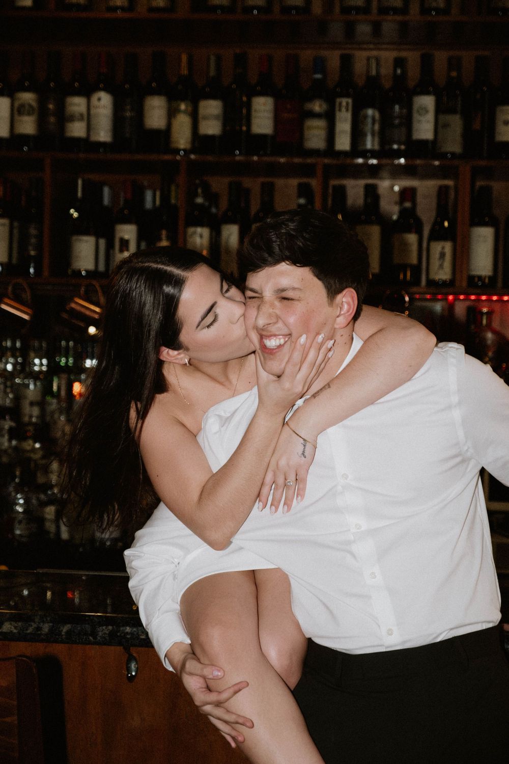 An intimate moment shared in a dimly lit bar setting with bottles in the background.