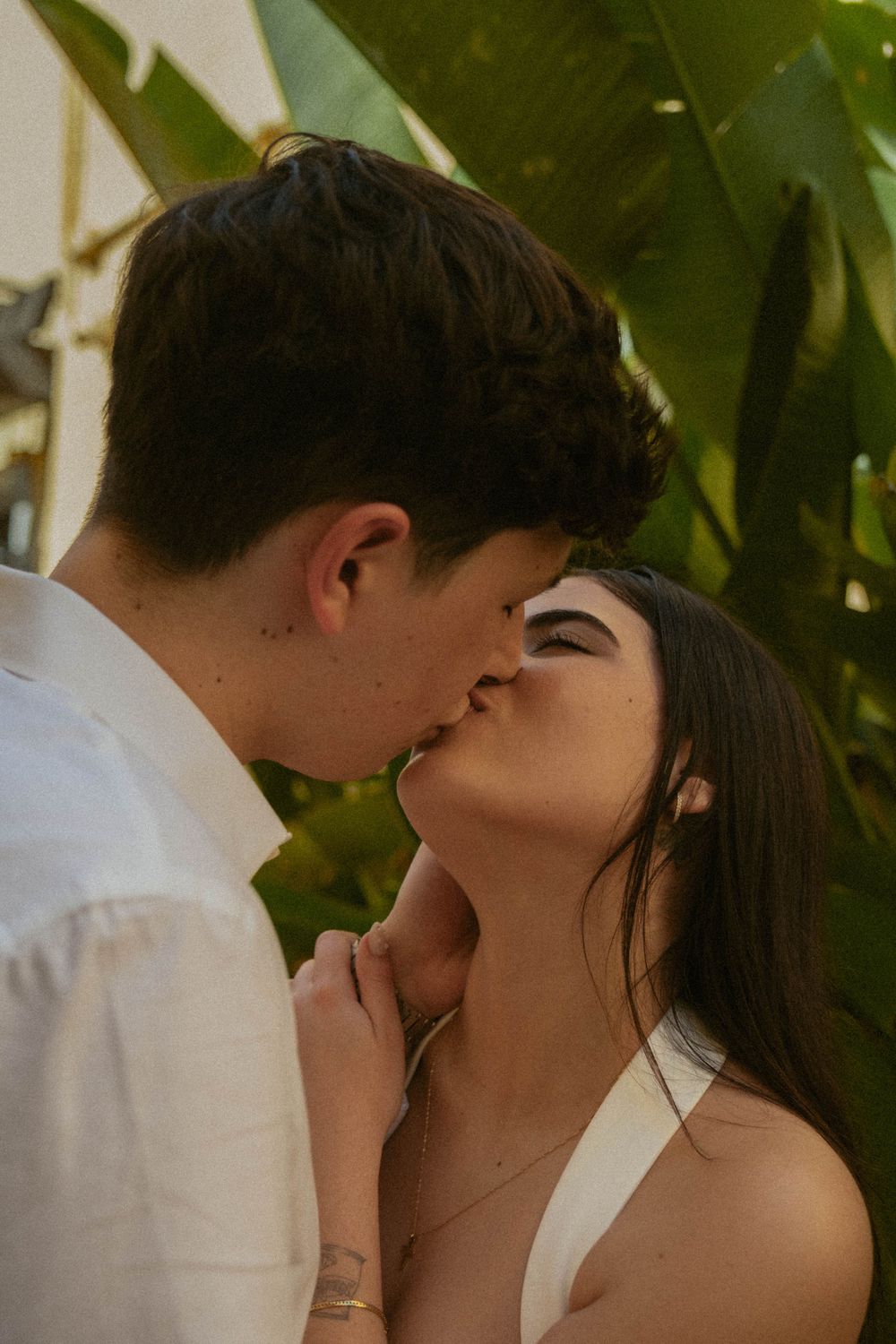 A romantic couple shares an intimate kiss outdoors against a backdrop of lush greenery.