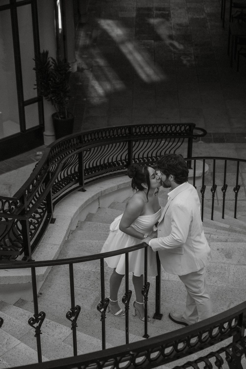 A black and white photo captures a romantic moment on a curved staircase with iron railings.