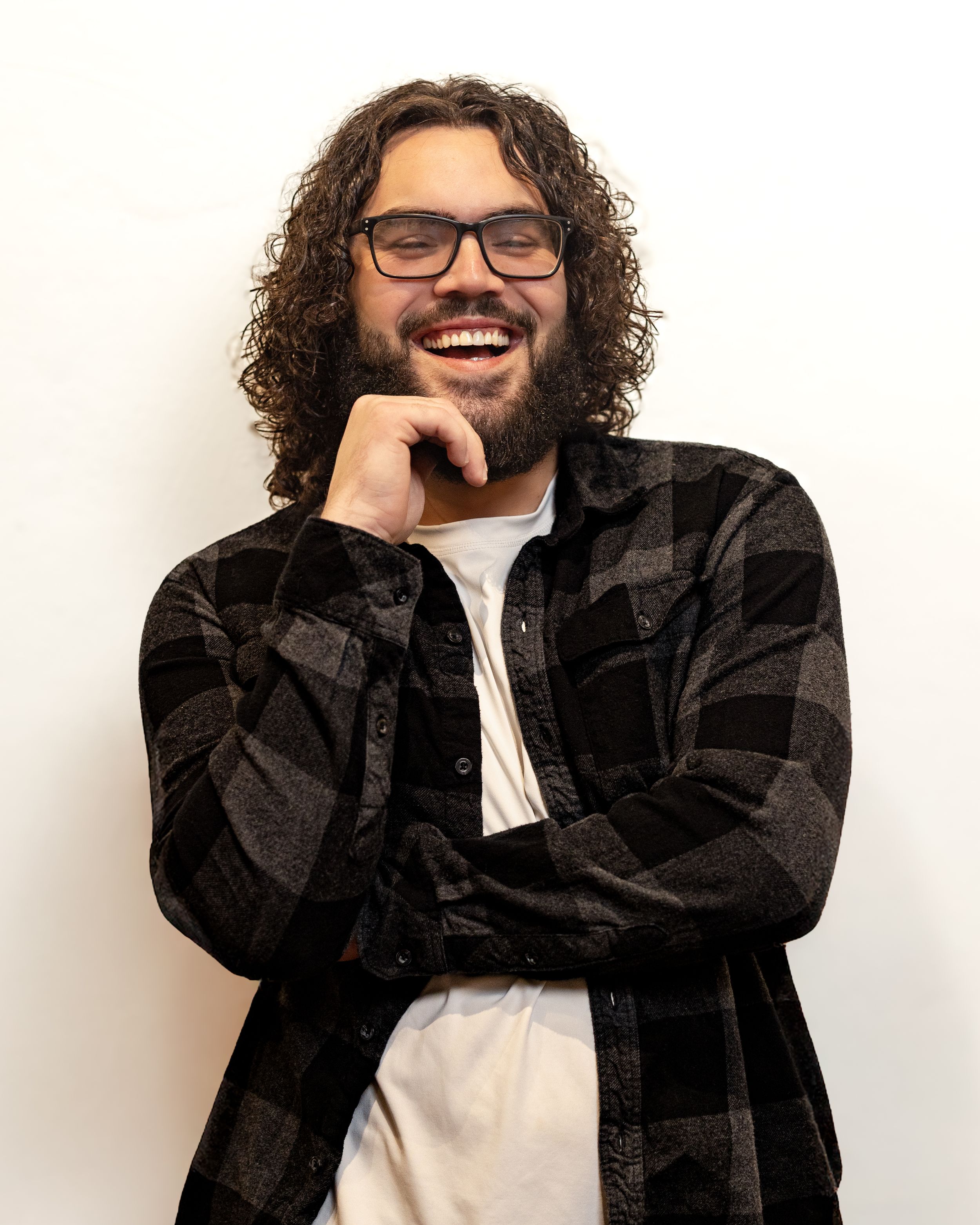 Smiling young man with curly hair, beard, and glasses wearing a plaid shirt, posing against a white background.