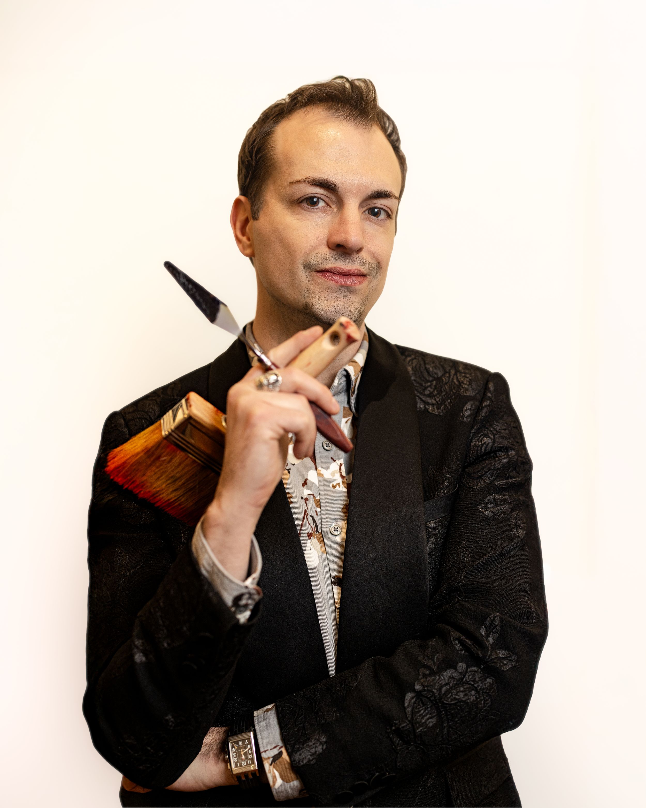 Elegant man in black brocade blazer holding a paintbrush and book, posing against white background.