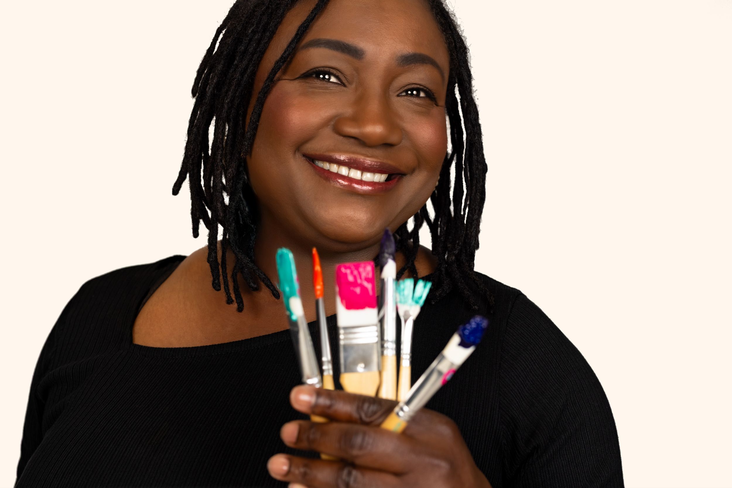Smiling woman holding colorful paintbrushes against a light background.