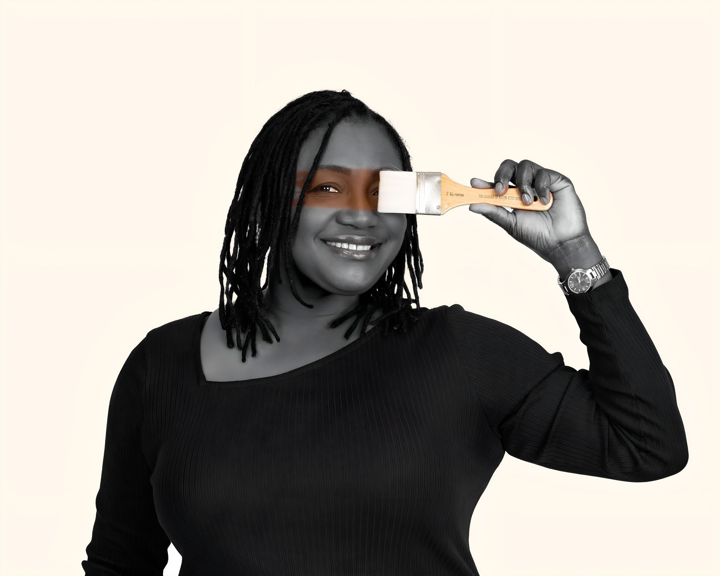 Smiling Black woman in black top holding a paint brush over one eye against cream background.