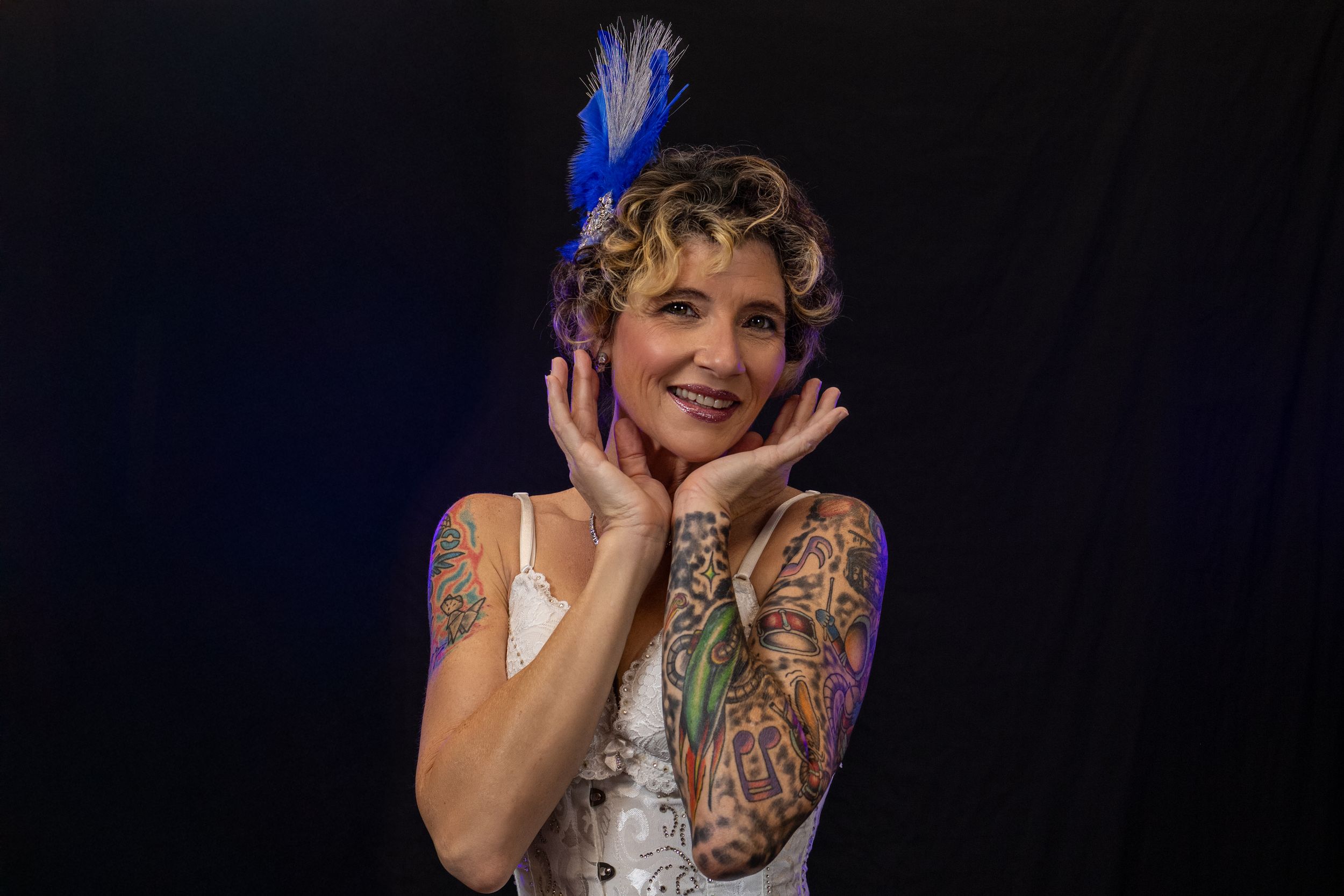Smiling woman with curly blonde hair, blue feather headpiece, and colorful tattoos on arms poses against dark background.