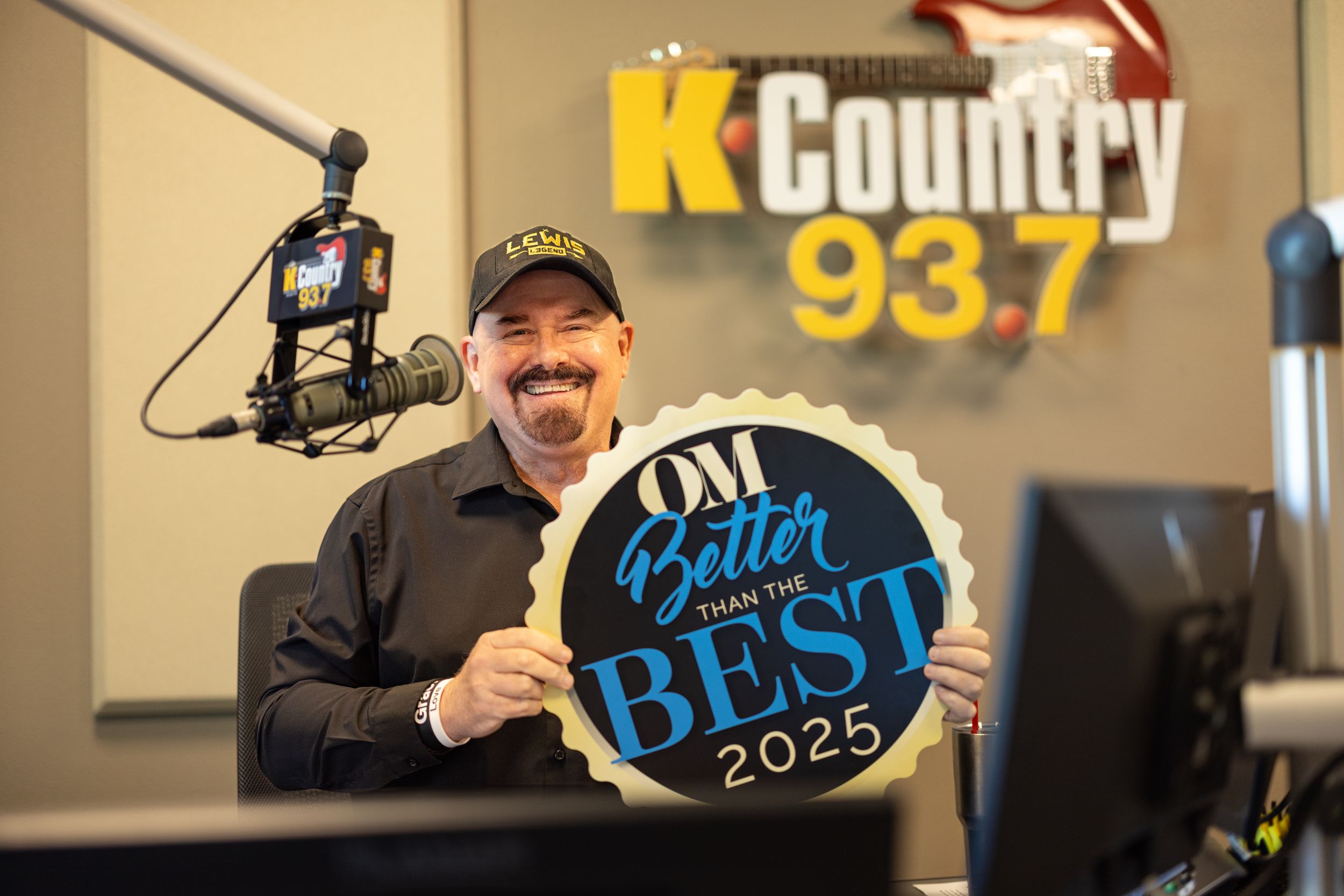A smiling radio host holds a Better Than the Best 2025 award at K Country 93.7 studio.