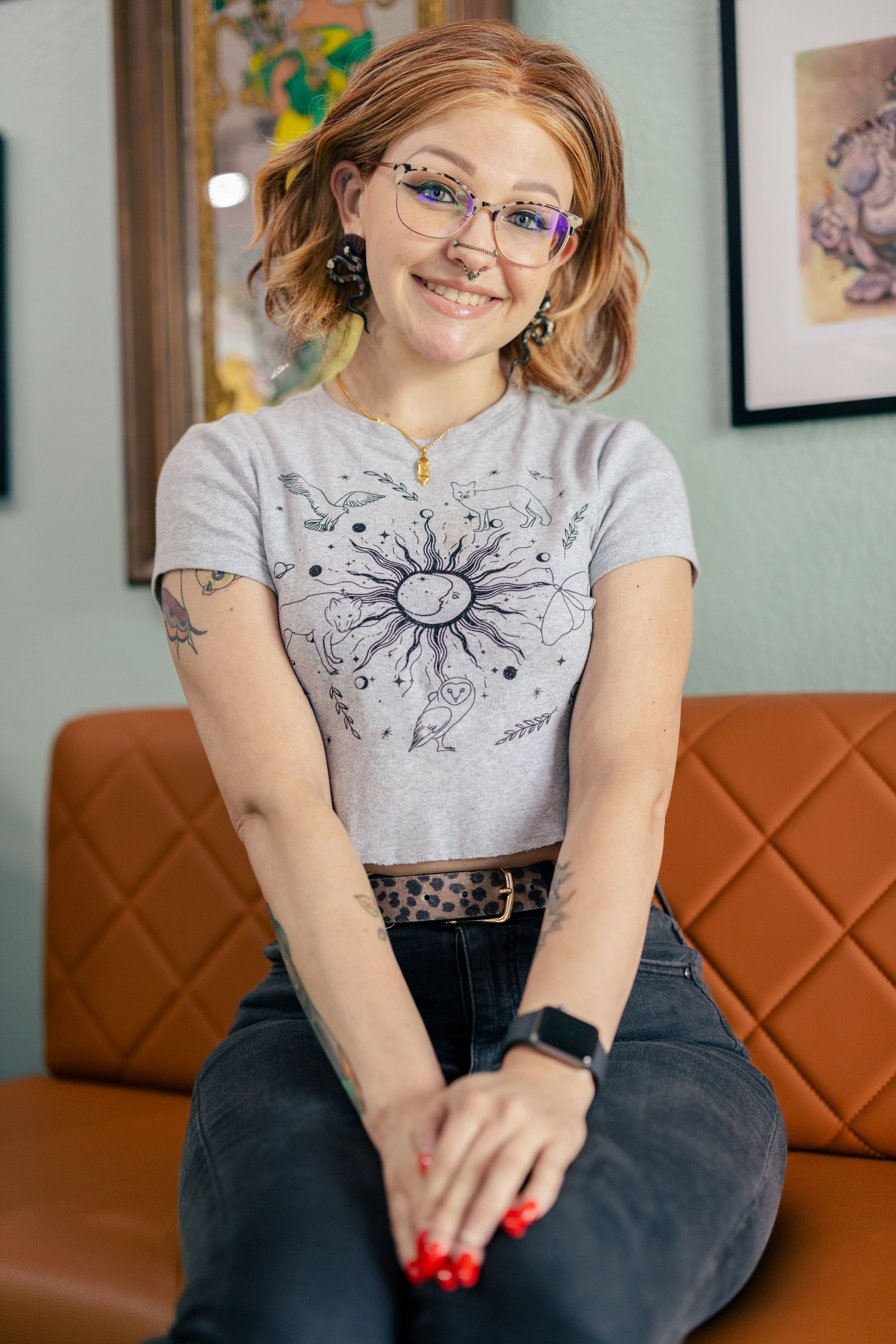 Smiling young woman with glasses and tattoos wearing a celestial sun graphic tee, seated on a brown leather couch.