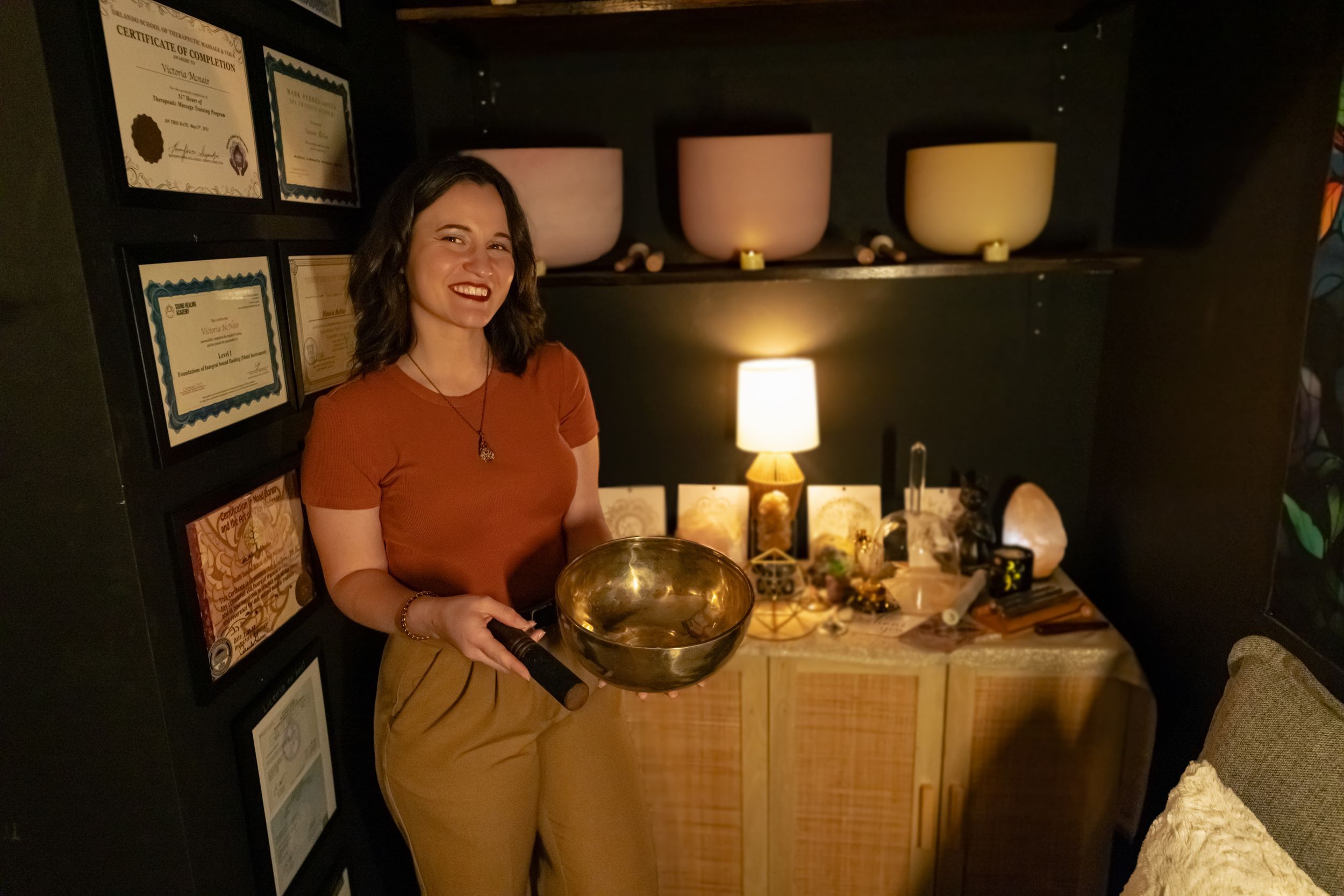 Smiling female therapist holding singing bowl surrounded by crystal singing bowls and holistic healing certificates.