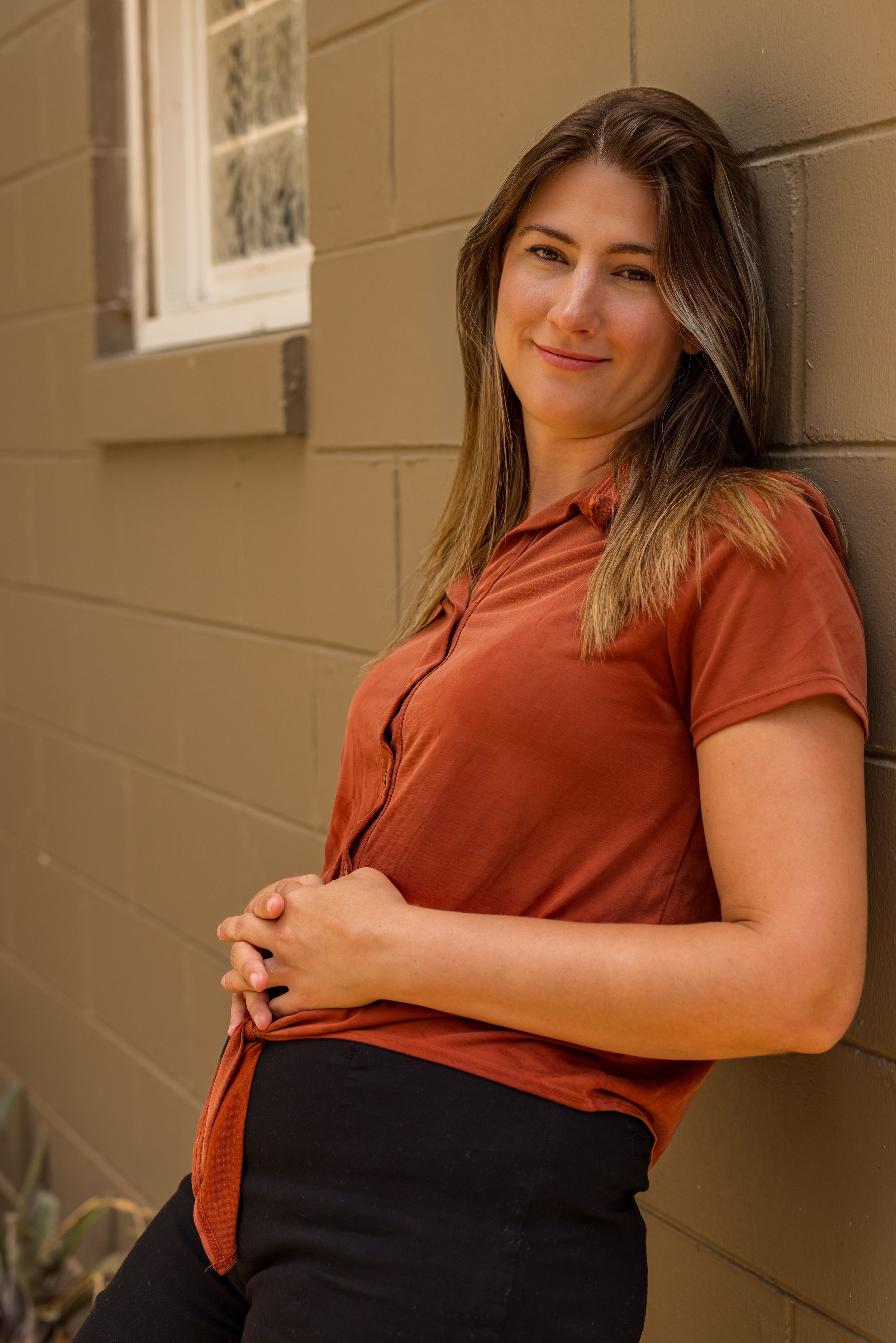 Young woman in rust-colored shirt leaning against a tan brick wall, smiling softly outdoors.