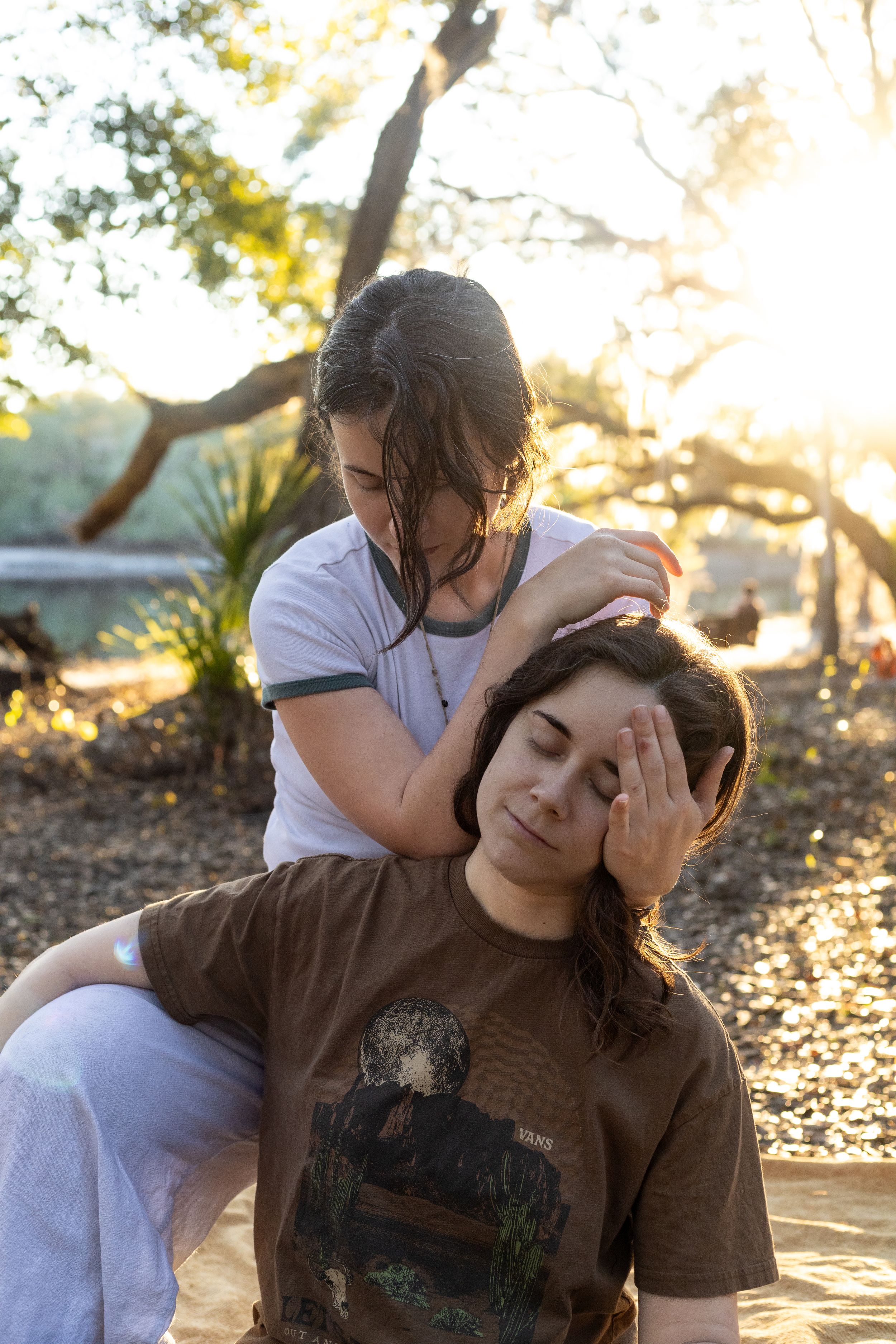 Two women outdoors in golden sunlight, one giving the other a relaxing head massage amid trees.