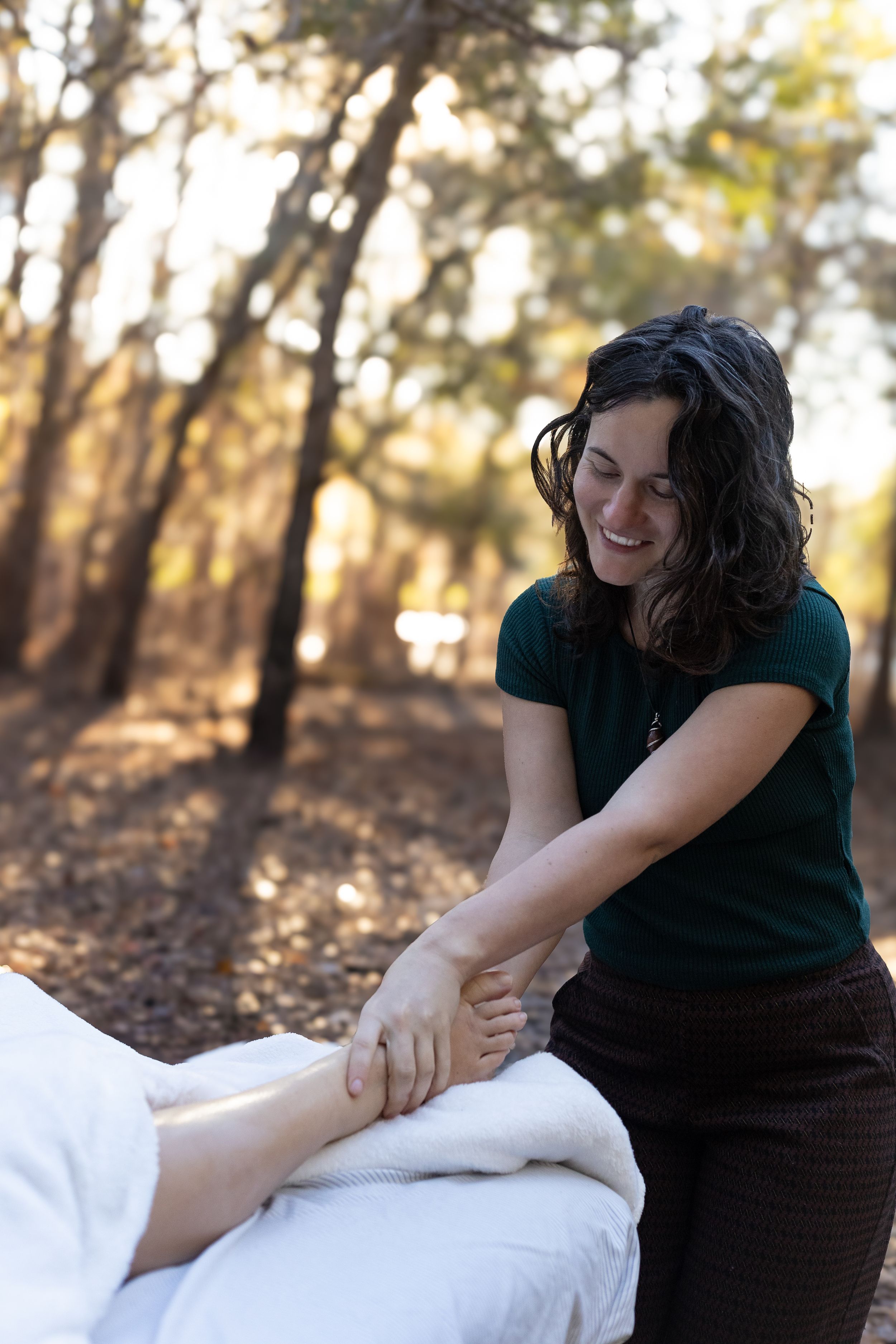 Smiling female massage therapist performing outdoor foot massage in a sunlit forest setting.