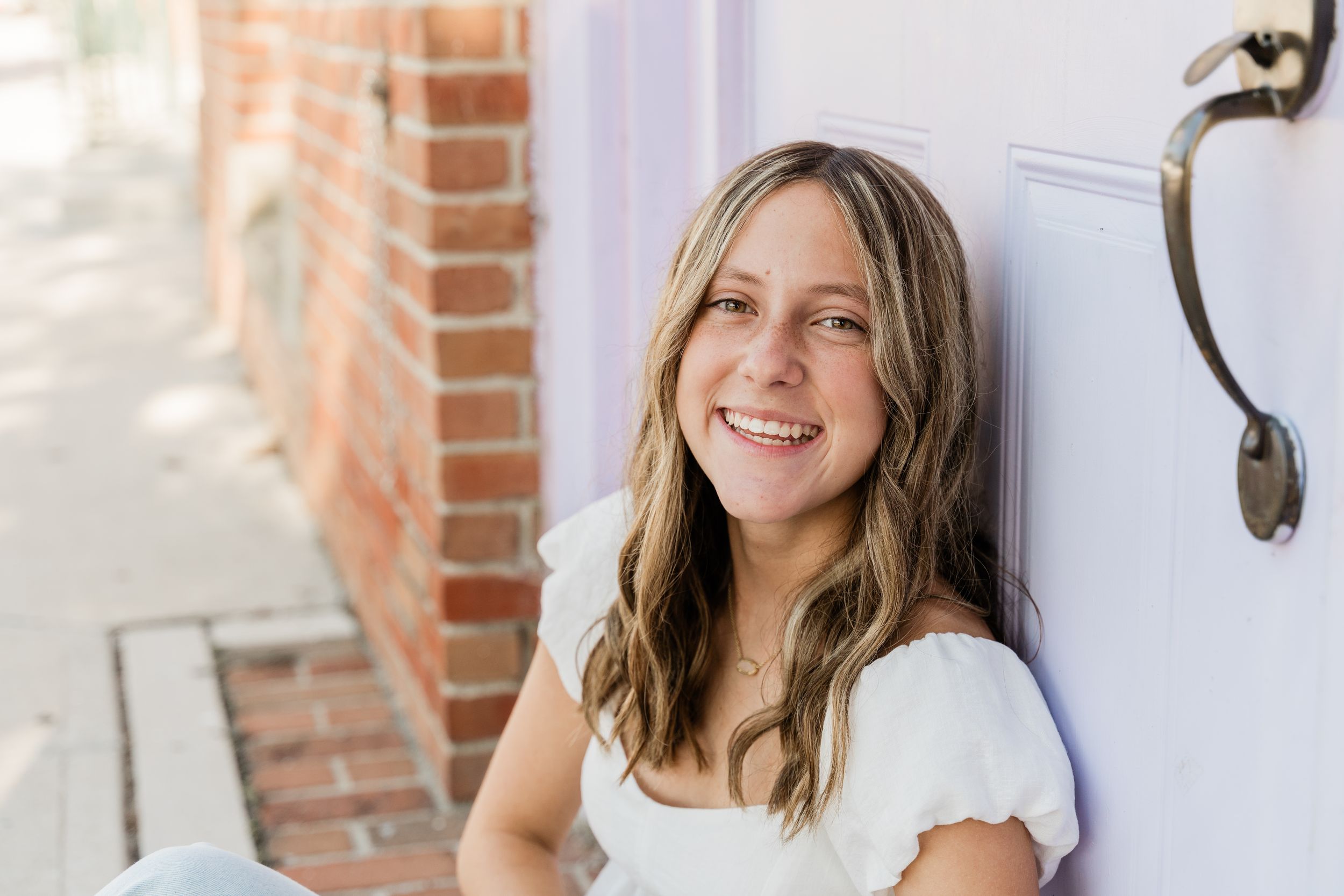 Woman in white top smiling and posing against brick wall in casual outdoor photo shoot.