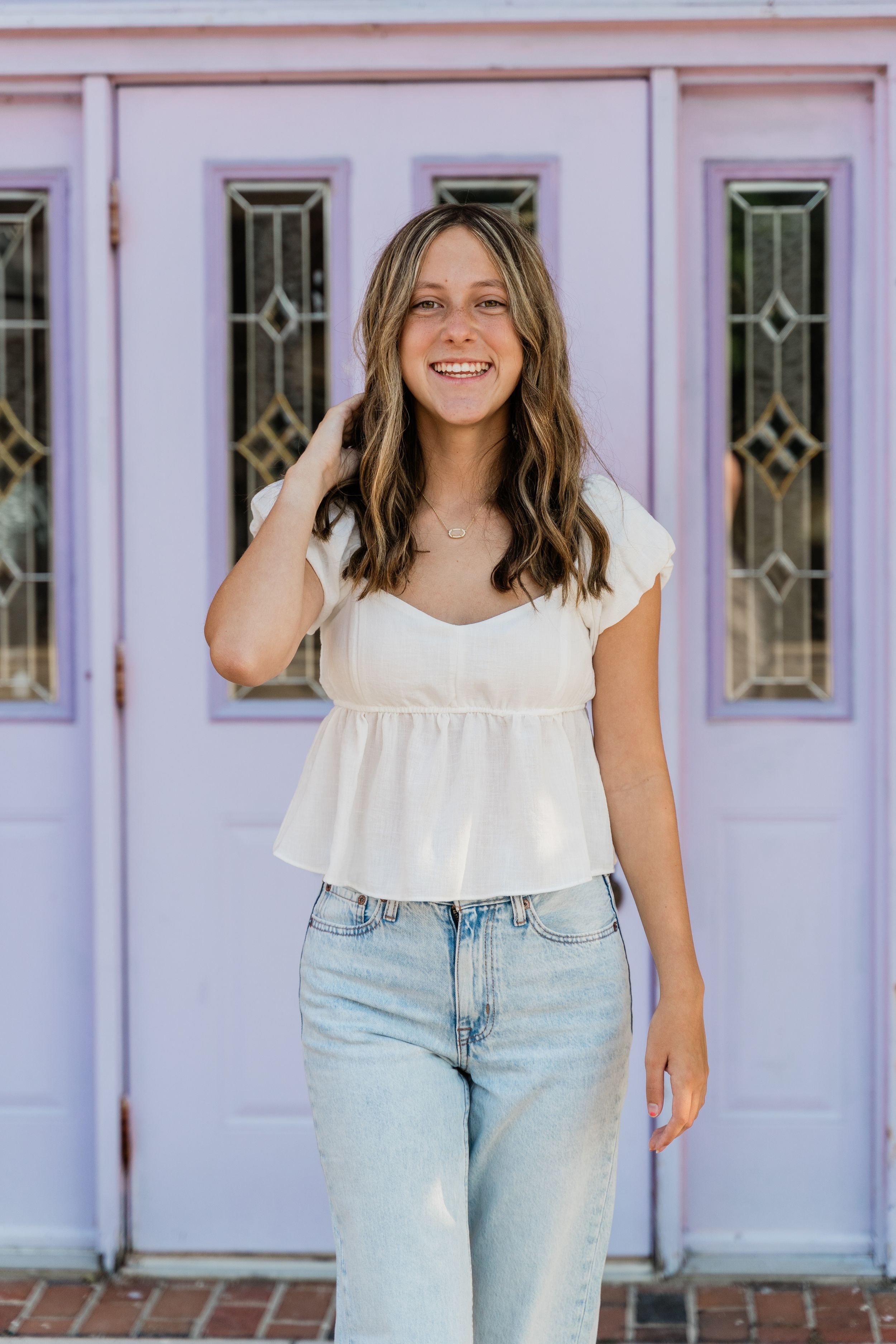 Three photos showing someone in a white ruffle top and light wash jeans posing against a pink building exterior.