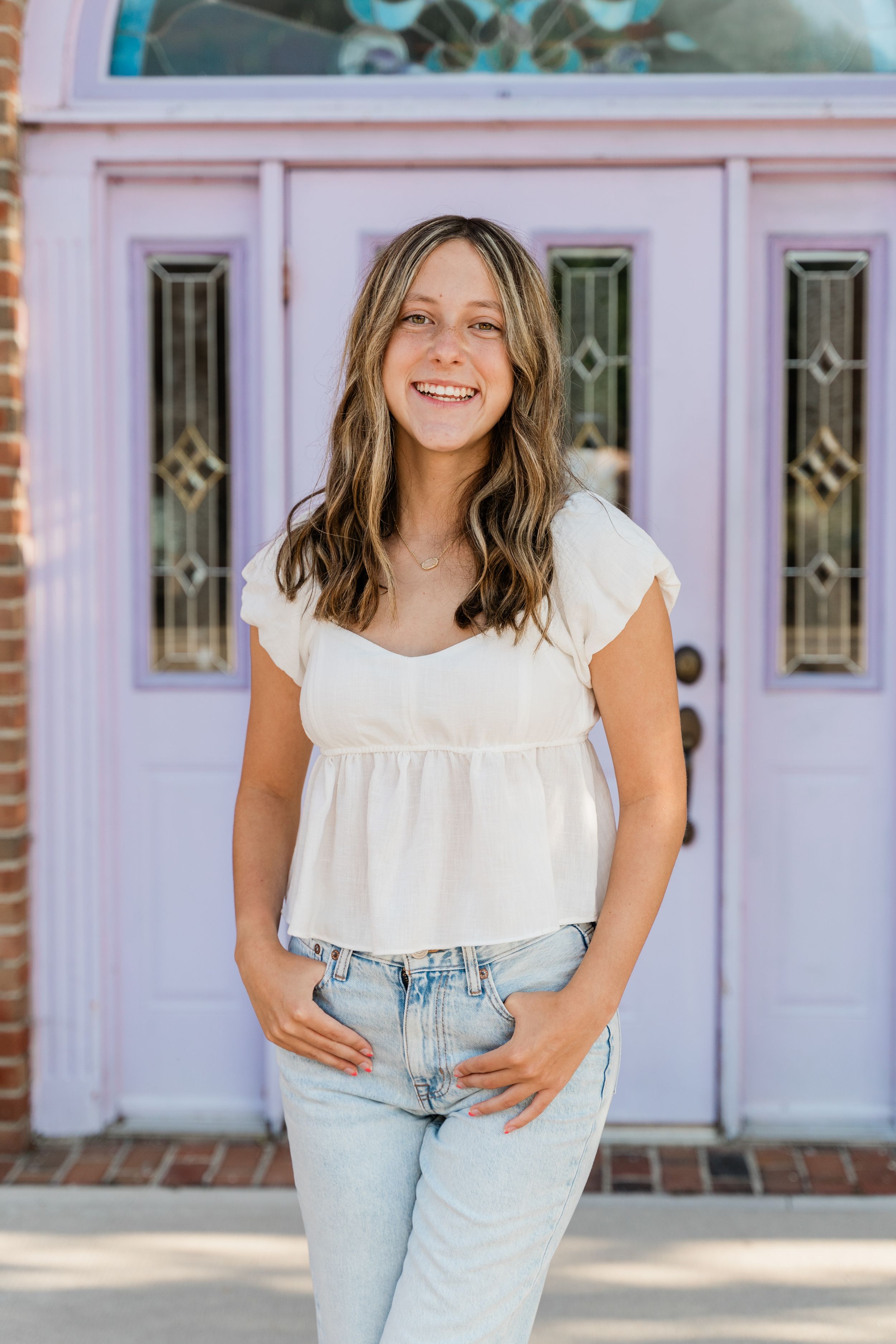 A person in a white babydoll top and light wash jeans stands smiling in front of a pink door with decorative glass panels.