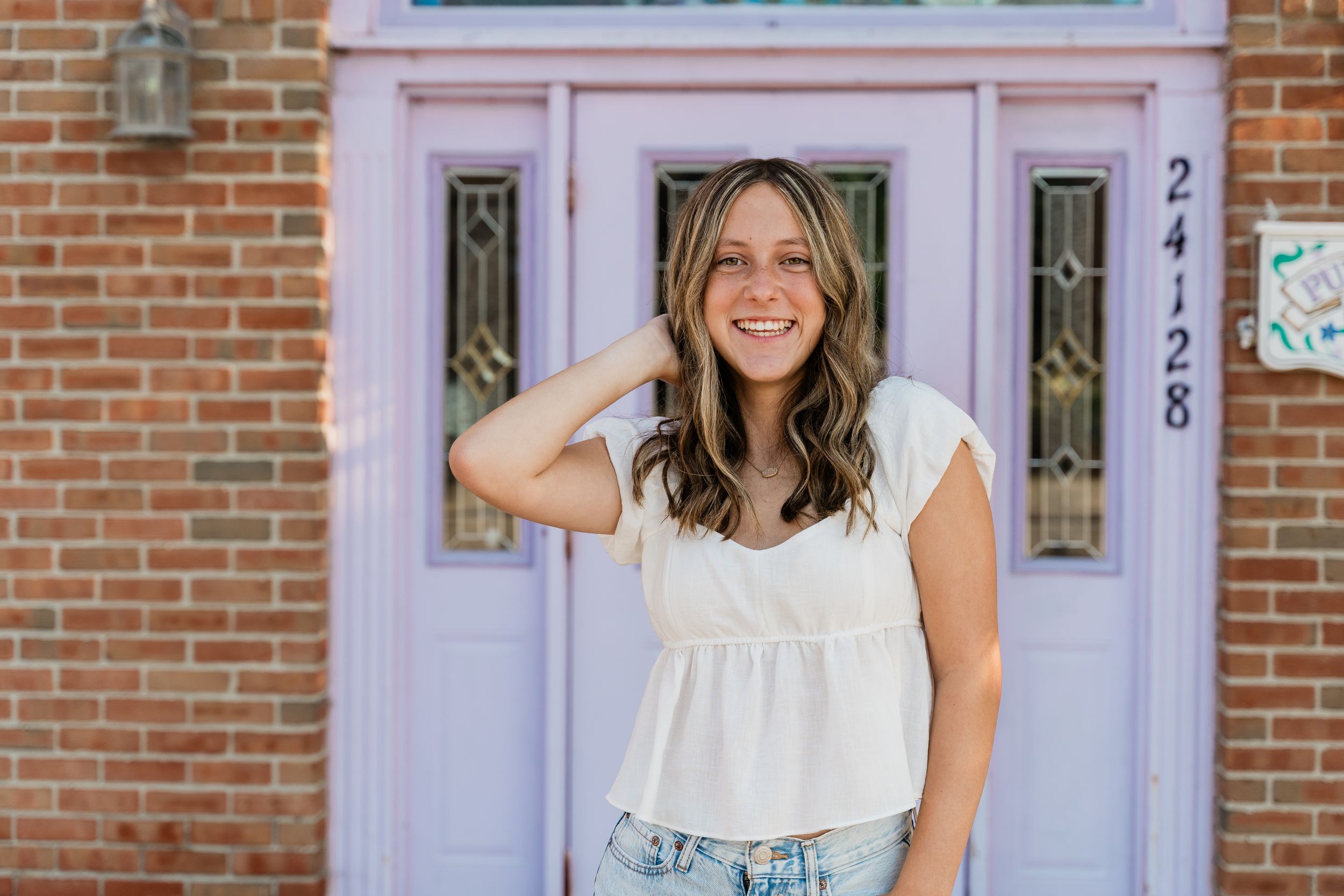 A woman in a white dress stands smiling in front of a brick wall and lavender-colored door.