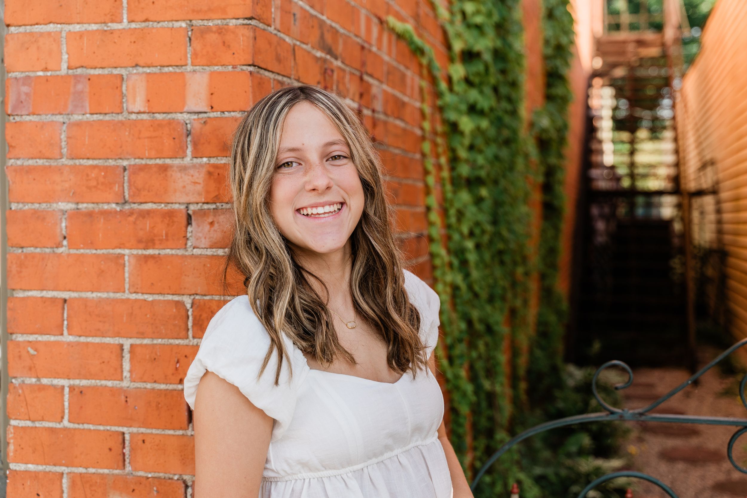A person in a white dress smiles while leaning against a red brick wall with ivy growing in the background.