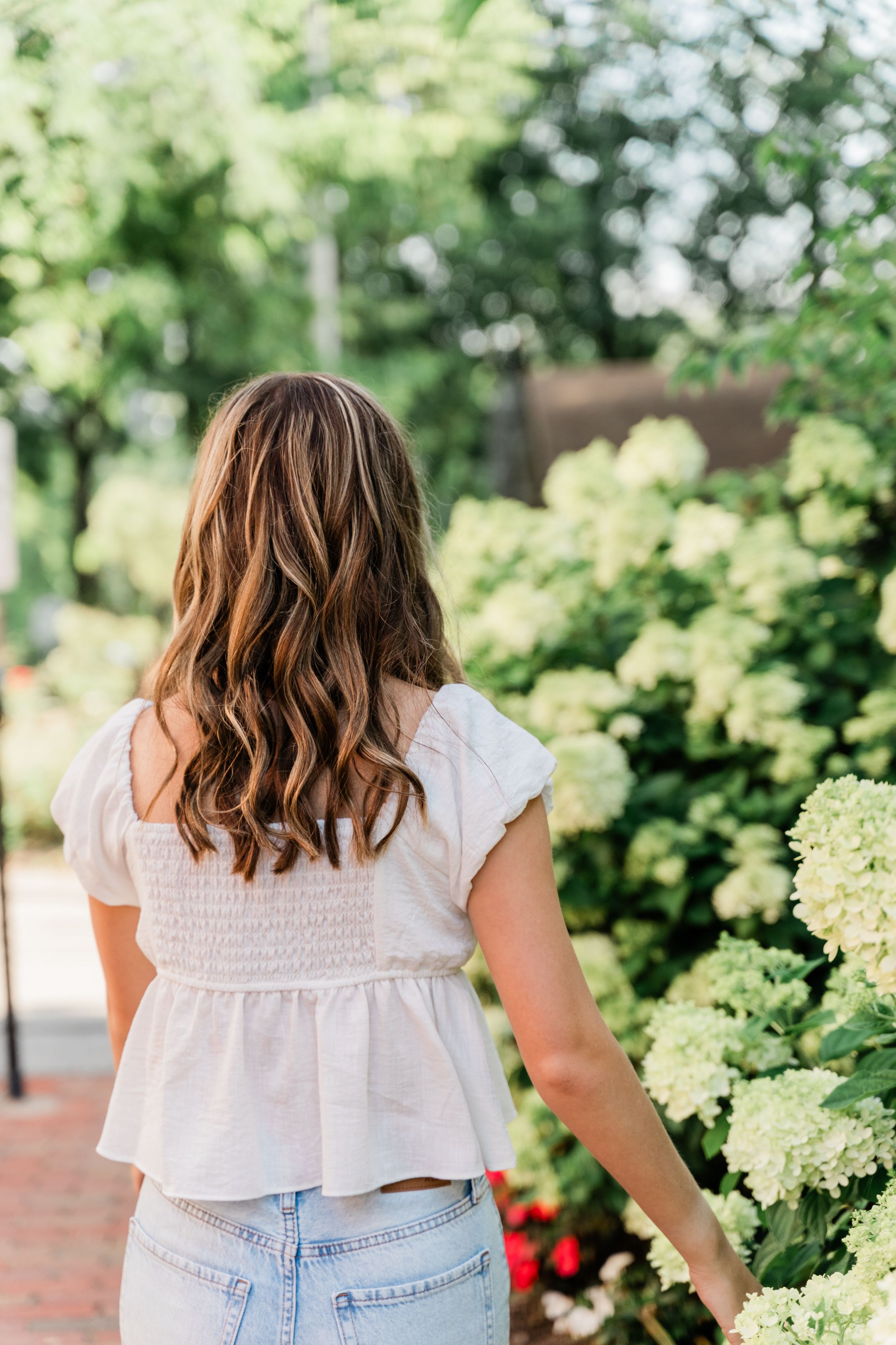 Person in white ruffled top and jeans stands near blooming hydrangea bushes in a garden setting.