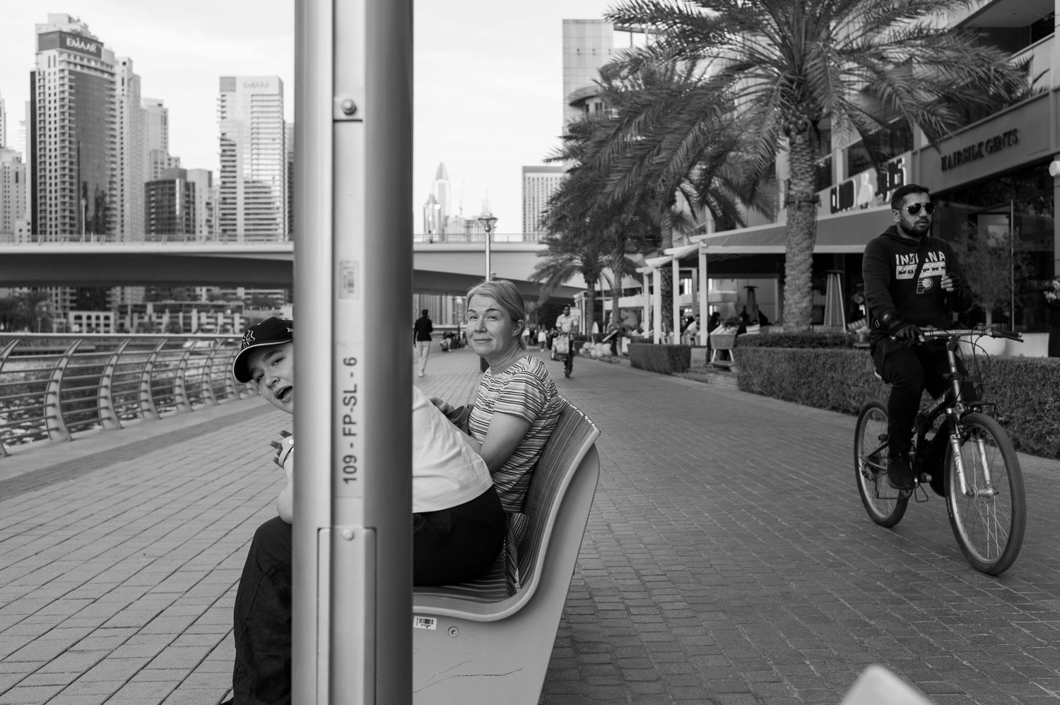Black and white urban scene of Dubai's waterfront promenade with palm trees, cyclists and city skyline in background.