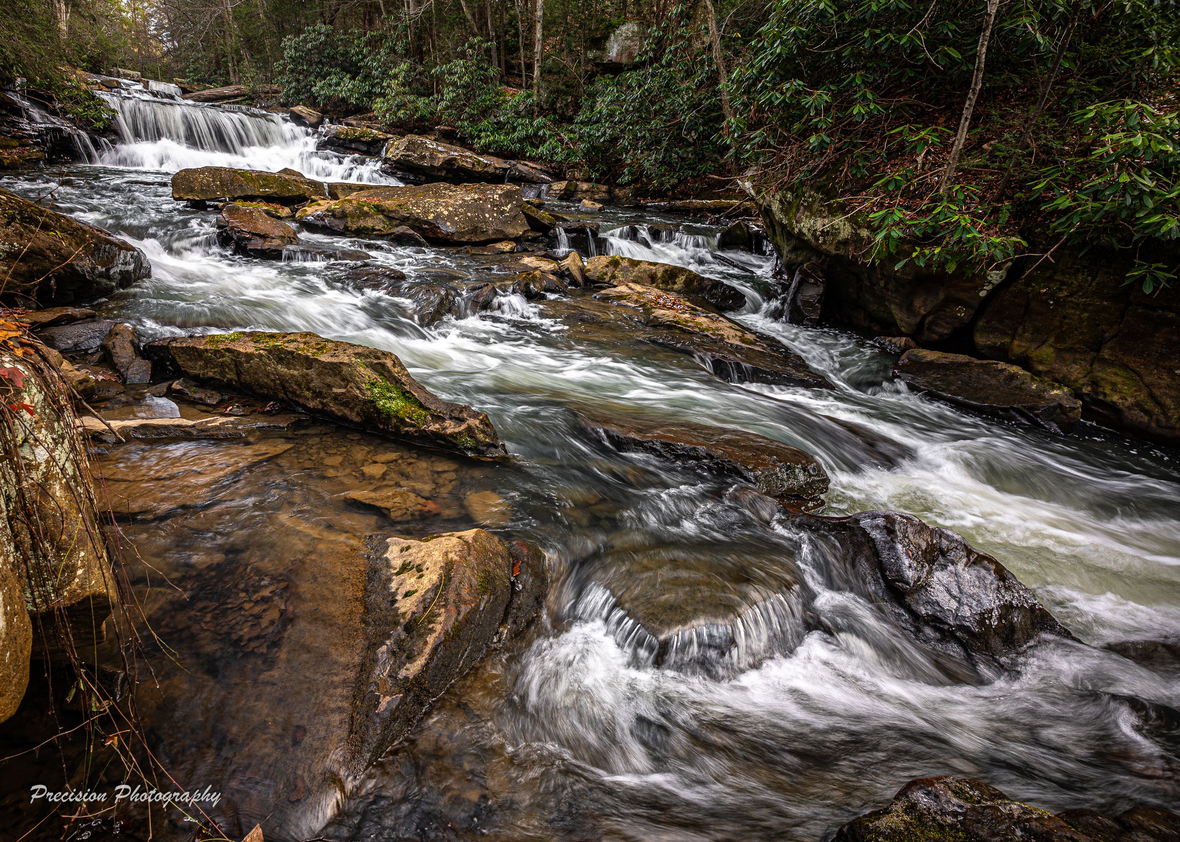 Multiple tiers of rushing waterfalls cascade over rocky terrain in a wooded landscape.