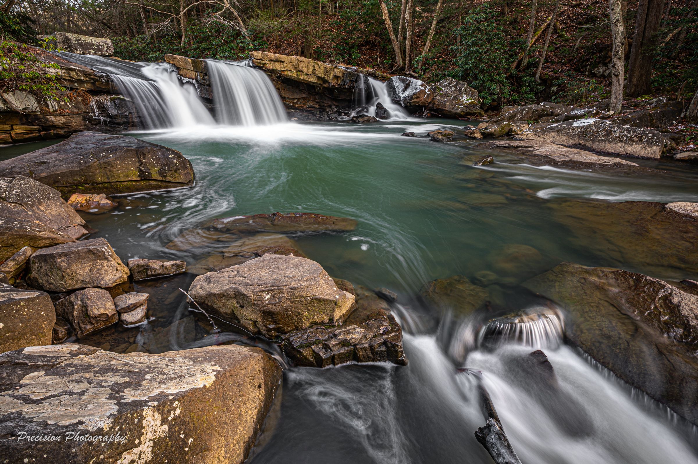 Scenic waterfall with turquoise pools flows through rocky forest terrain.