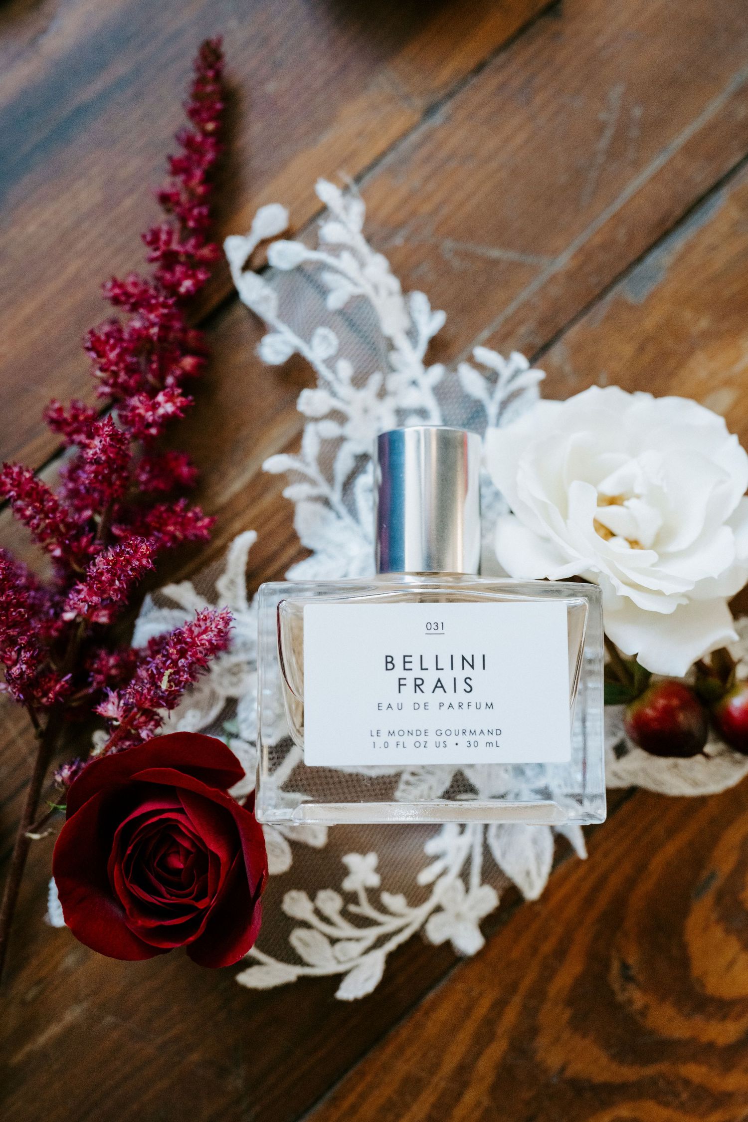 Perfume bottle labeled 'Bellini Frais' styled with white and burgundy flowers on rustic wooden surface.