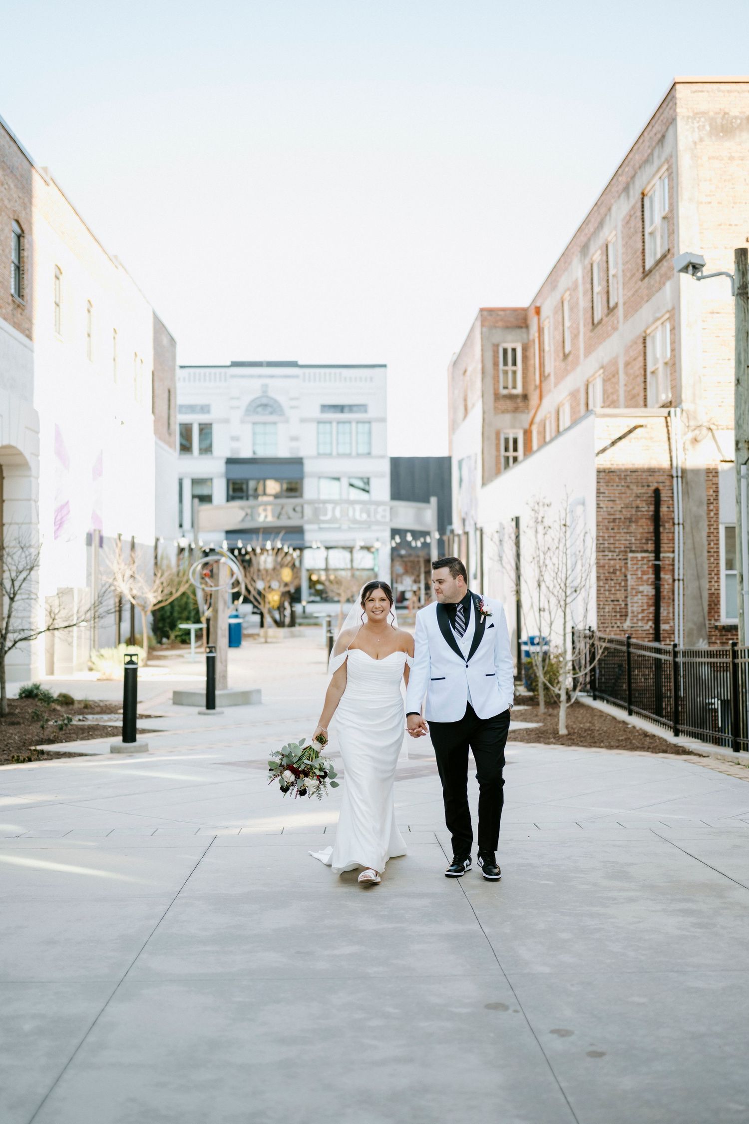 A couple walks together on an urban sidewalk, the bride in a white gown and groom in a white tuxedo jacket.