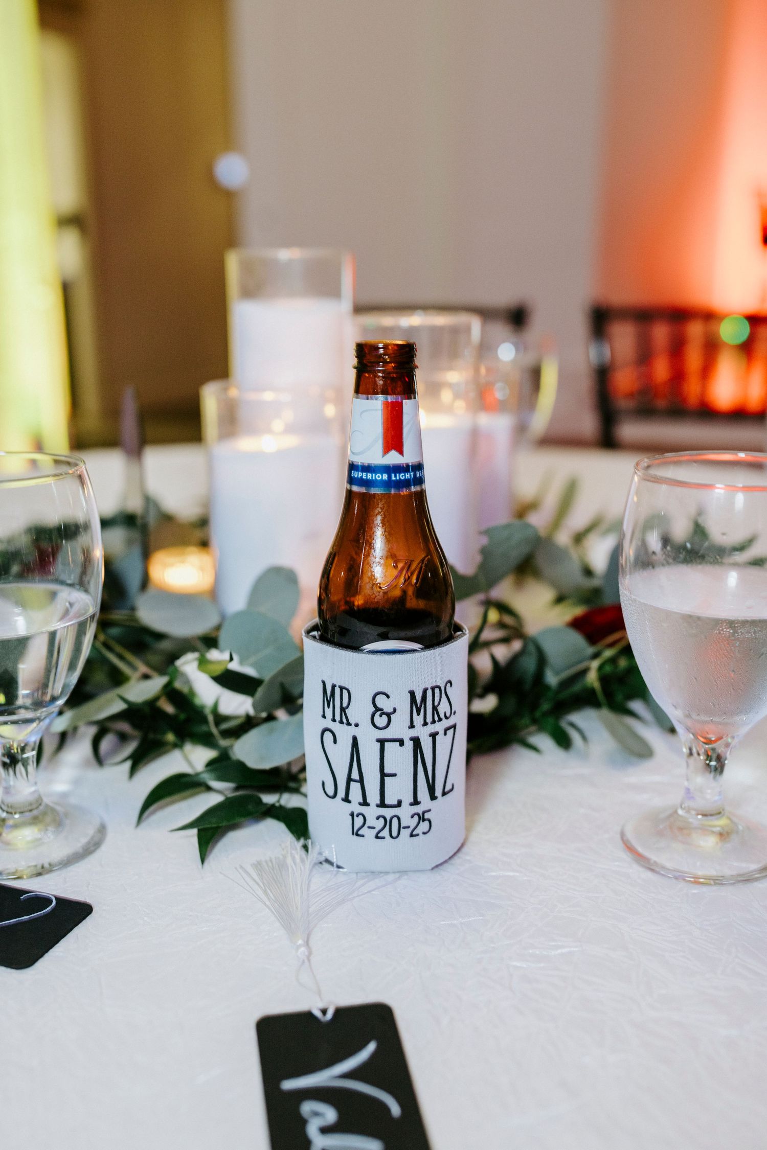 Wedding reception table decor features lit candles, greenery garlands, and personalized name cards on a white tablecloth.