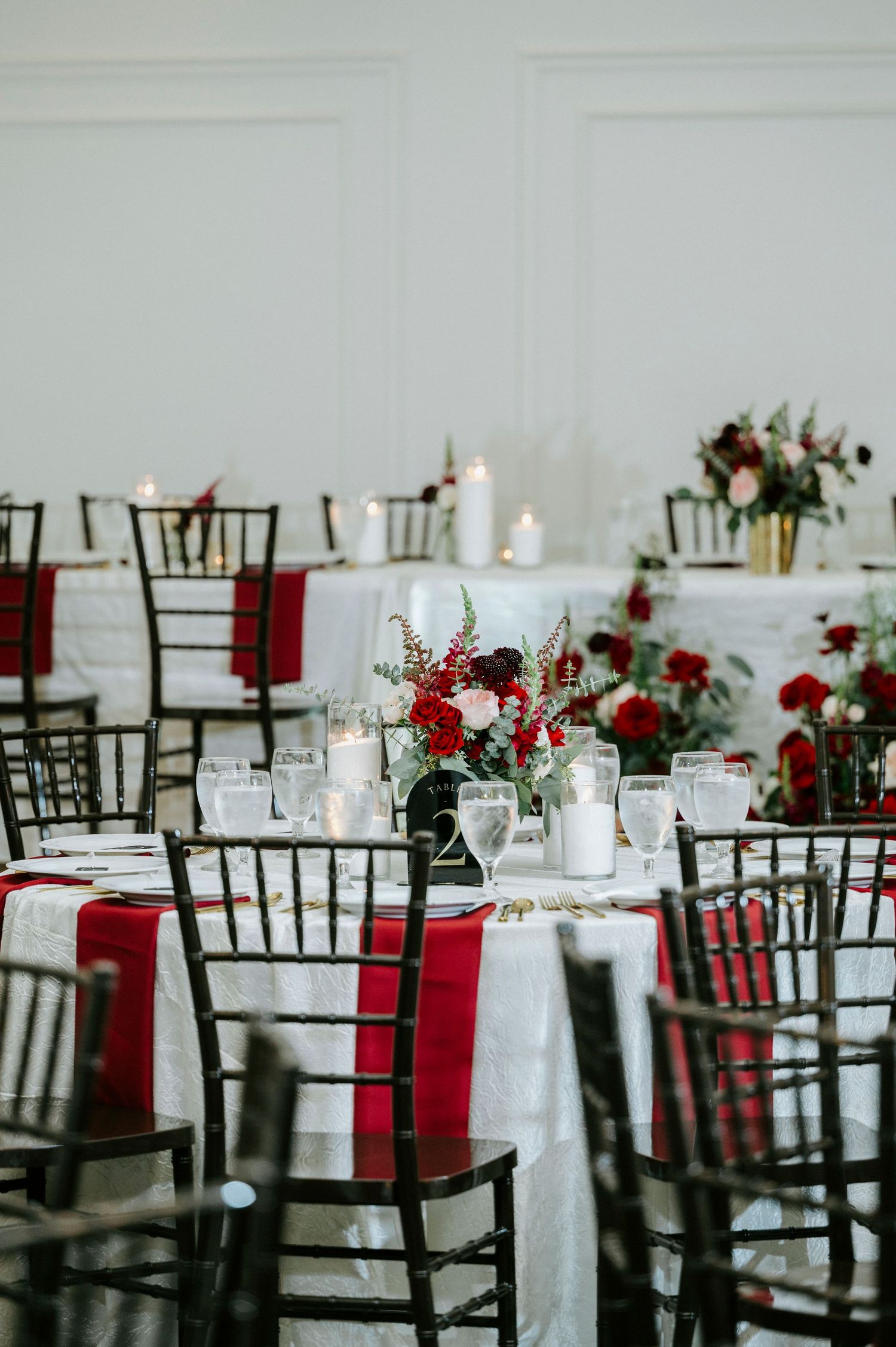 Elegant wedding reception tables decorated with dark chiavari chairs, white linens, red accents and romantic candlelight.