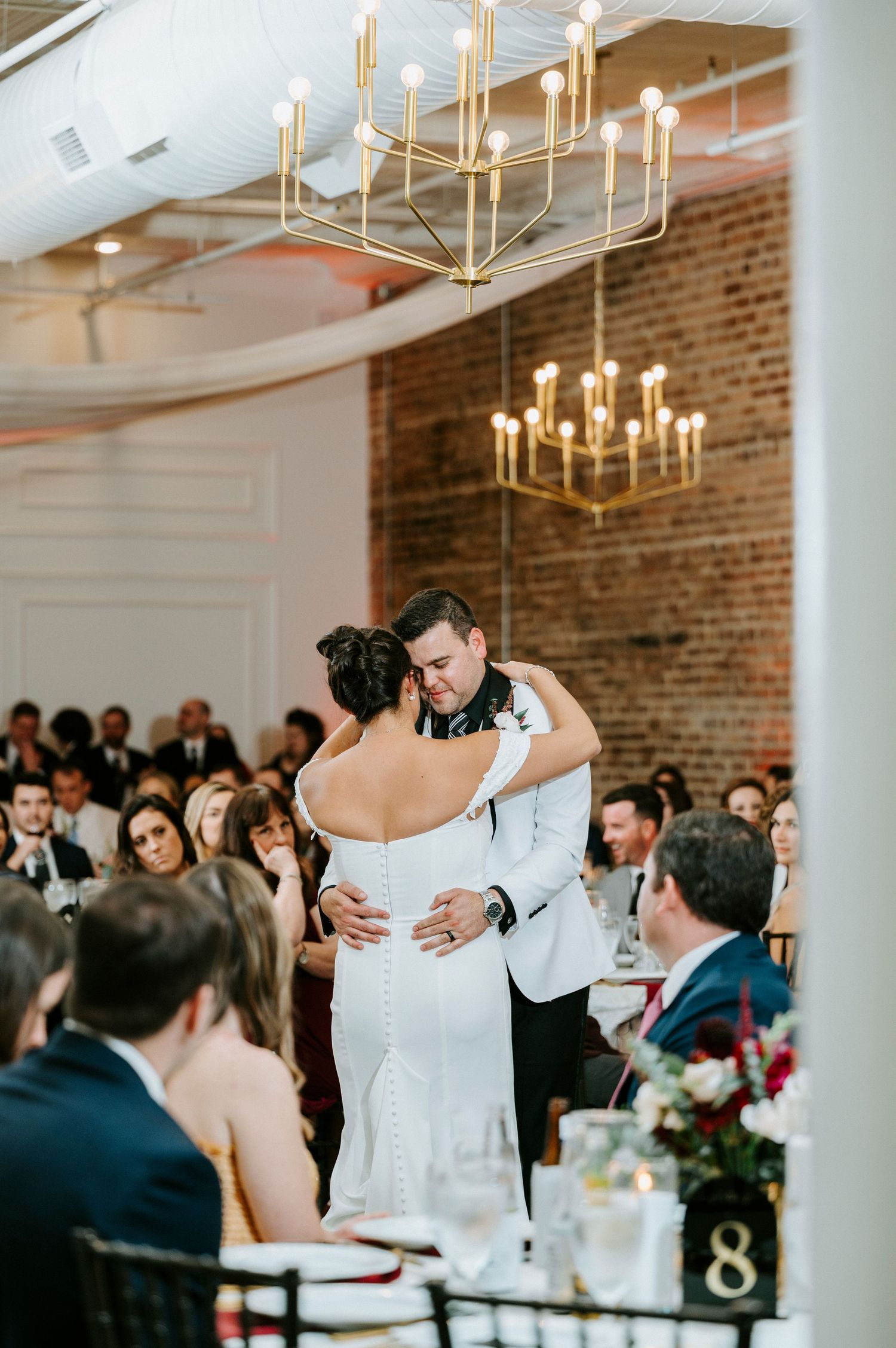 Couple sharing their first dance at a modern reception venue with exposed brick walls and gold chandeliers.