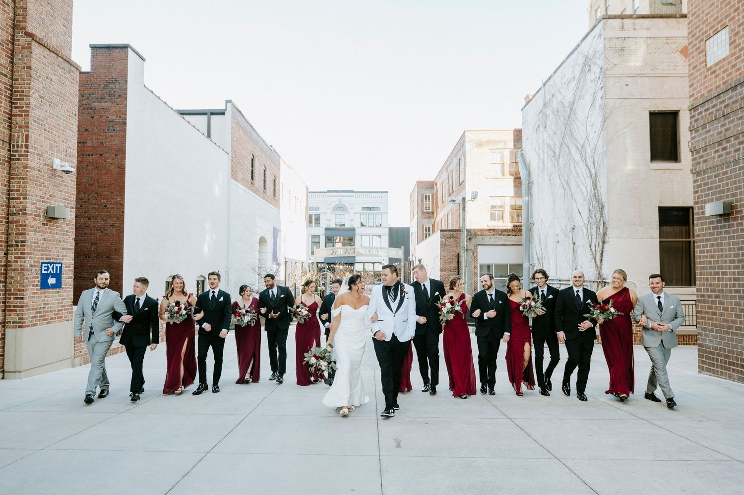 Wedding party in burgundy and black attire poses against snowy winter backdrop at outdoor venue.