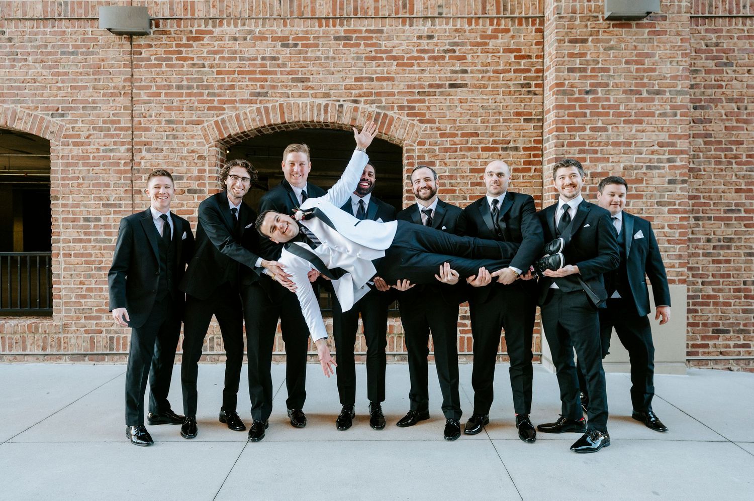 A group in formal black attire poses playfully in front of a brick wall in a series of candid shots.