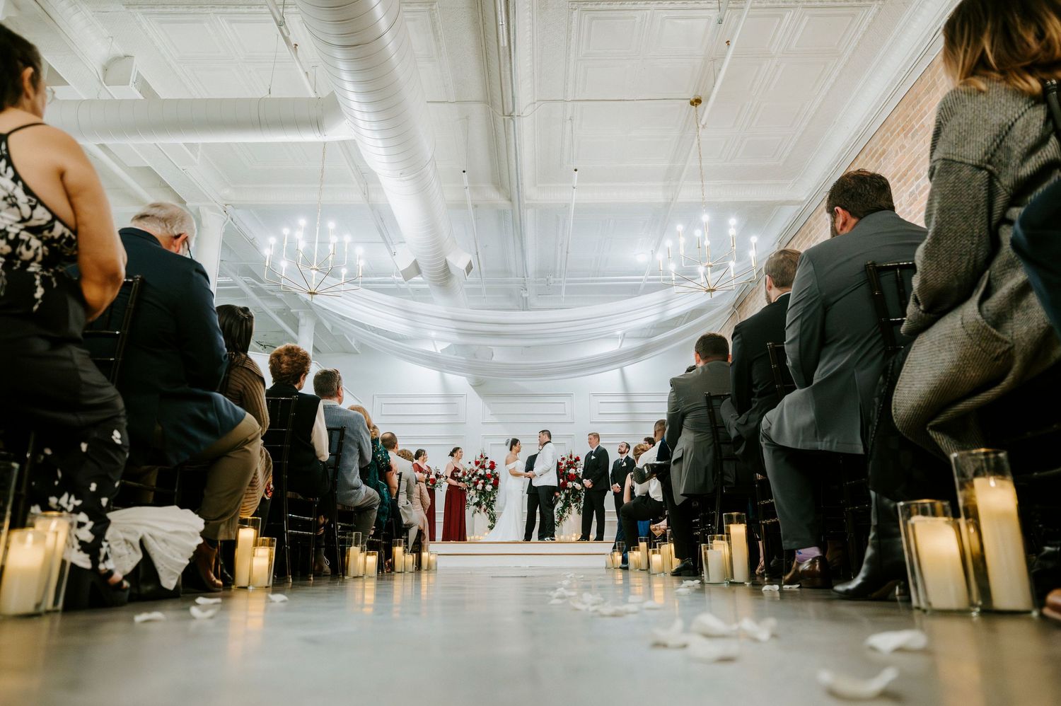 People in formal attire mingle at an elegant event with white draped decor and chandeliers overhead.