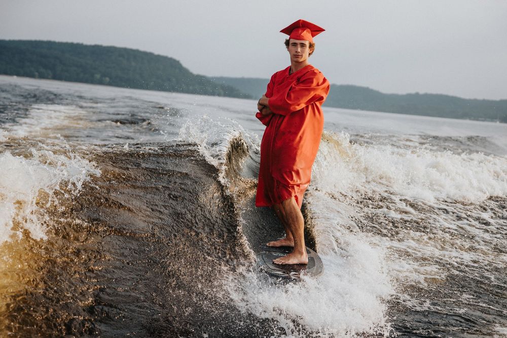 Graduate in red cap and gown surfing on choppy lake waters with mountains in background.