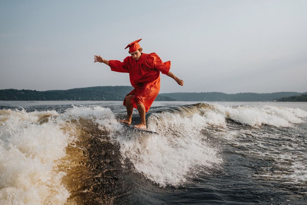 Person in red graduation gown performs acrobatic flips over ocean waves at sunset.