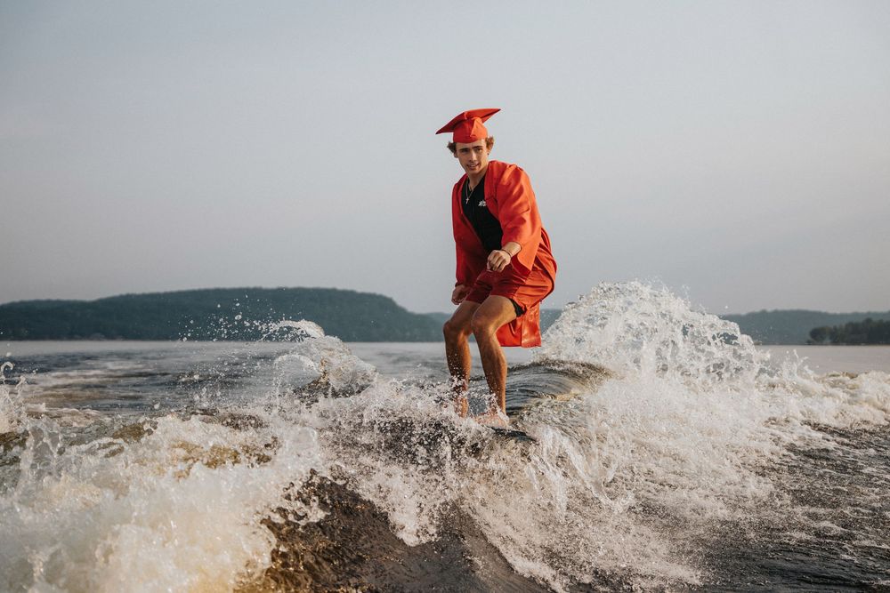 Person in graduation attire wakesurfing through splashing waves on a lake.