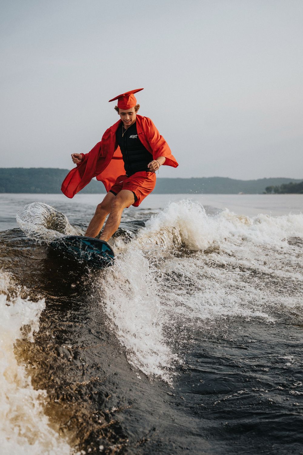 Graduate in red cap and gown surfing on a wave while celebrating graduation in a unique and adventurous way.