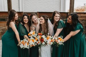 Bridesmaids in emerald green dresses holding orange and white bouquets pose together.