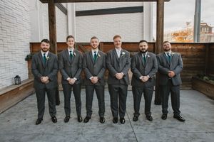 Groomsmen in grey suits sit lined up against white brick wall.