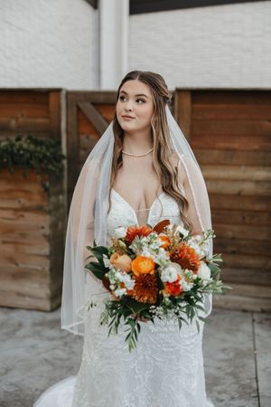 Bride in white lace dress holding vibrant orange and white fall bouquet.