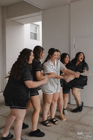 Bridesmaids in black dresses pose playfully against white wall.