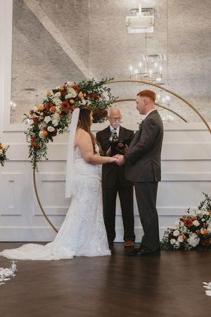 Wedding ceremony in progress with circular arch decorated with orange and white floral arrangements in elegant venue.