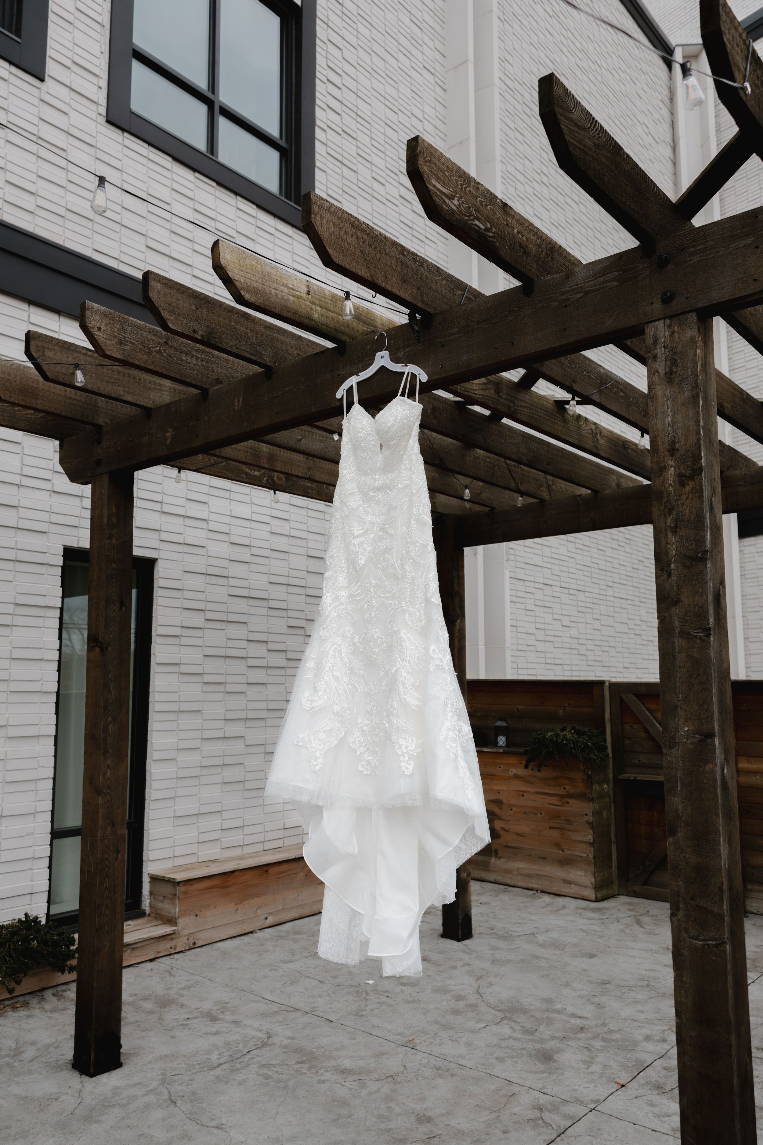 White lace wedding dress hanging from rustic wooden beams against a white brick wall.