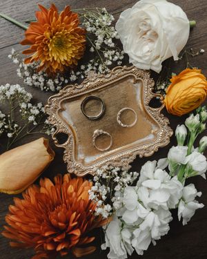 Wedding rings displayed on vintage silver tray with orange dahlias, white roses, and baby's breath flowers.