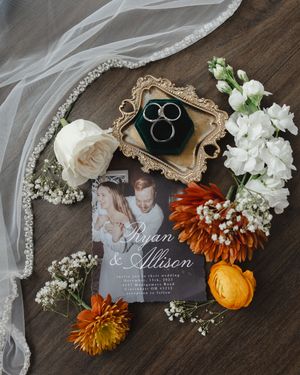 Wedding details flatlay with invitation, rings, flowers, and fall decorations on dark wooden surface.
