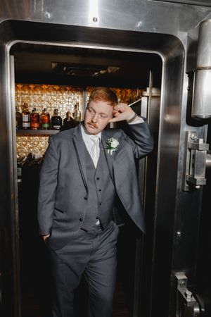 Groom in grey suit posing against stainless steel wall with bar glasses in background.