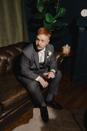 Wedding day portrait of groom sitting on brown leather couch in formal grey suit.