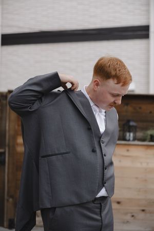 Getting ready photos of a groom in a classic charcoal suit against wooden wall backdrop.